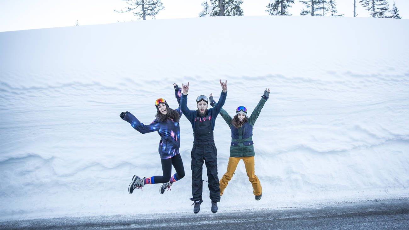 Three women jump up in front of a huge berm of snow.