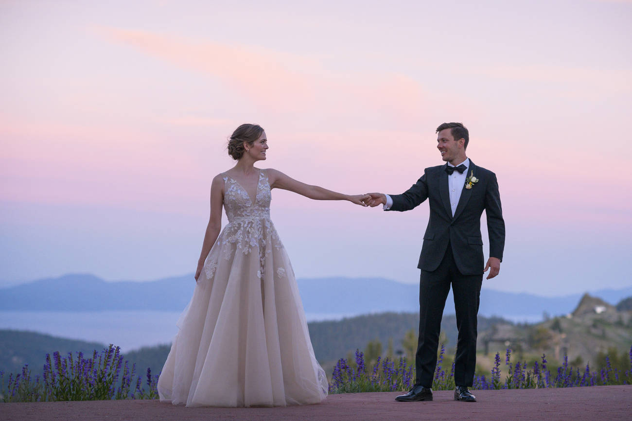 Bride and groom hold hands with lupines and Lake Tahoe in the background at Squaw Valley during a summer sunset