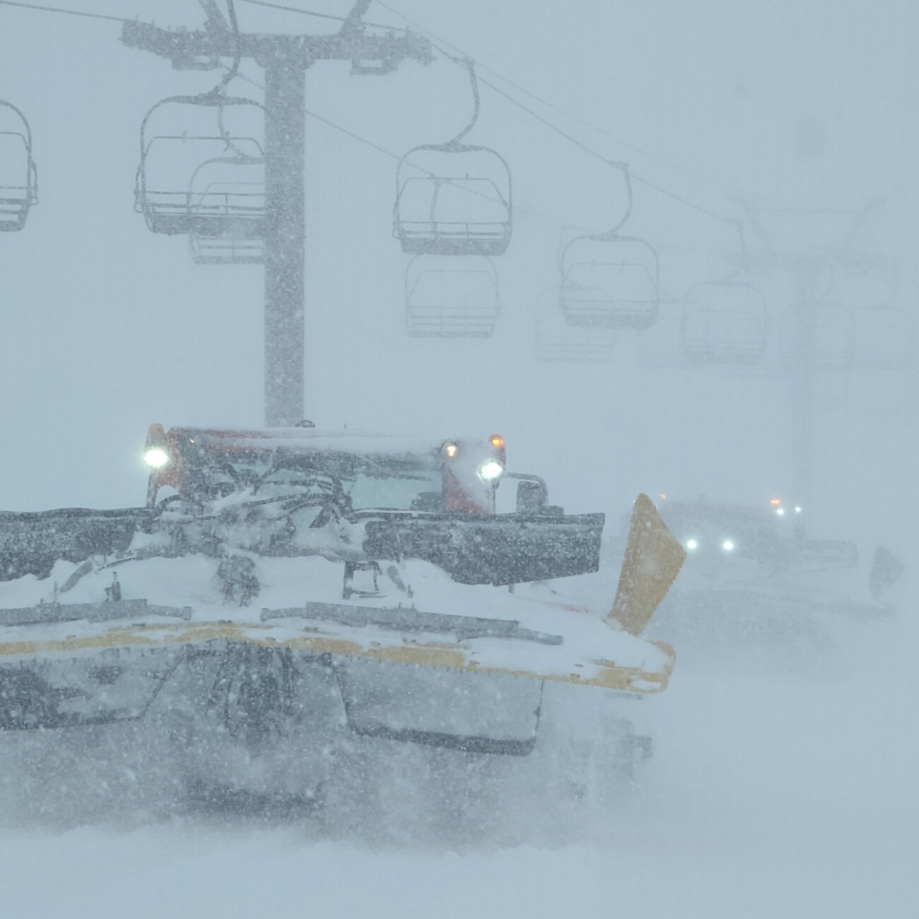Snowcats crossing under a chairlift in heavy snowfall.
