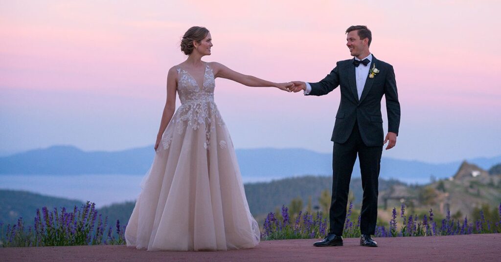 Bride and groom stand in front of pink and purple sunset at Palisades Tahoe with view of Lake Tahoe in background.