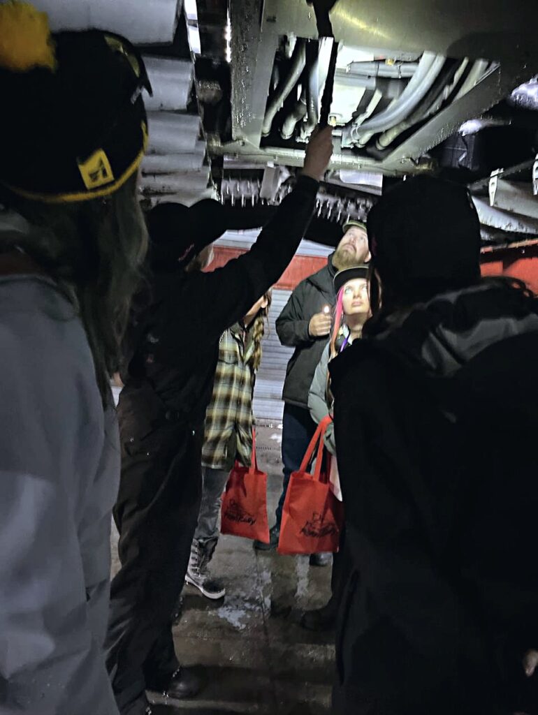 SheGrooms participants standing beneath a snowcat in the vehicle shop.